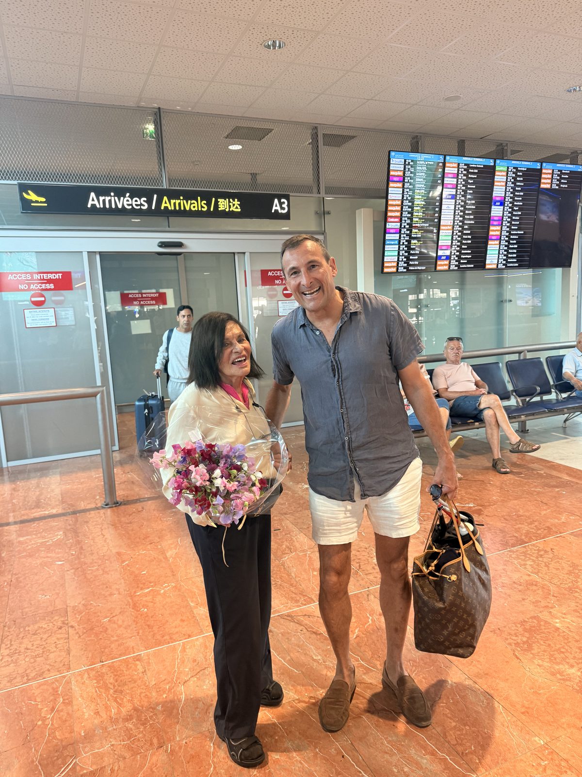 Two of the family meeting in the arrivals hall at Nice airport, one holding a bouquet