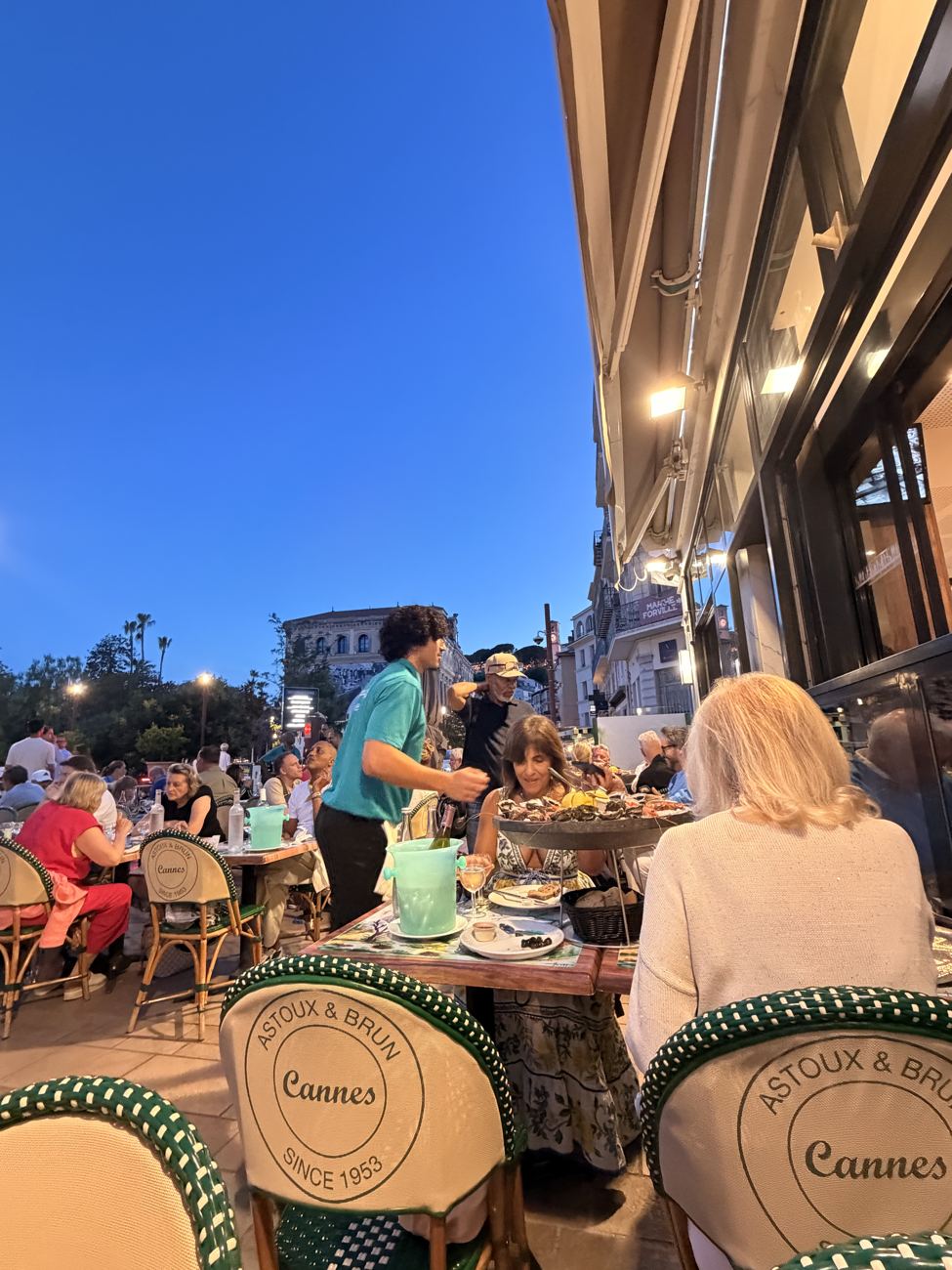 A crowded Cannes café terrace at blue hour, awnings and lamps lit, the deep blue evening sky above