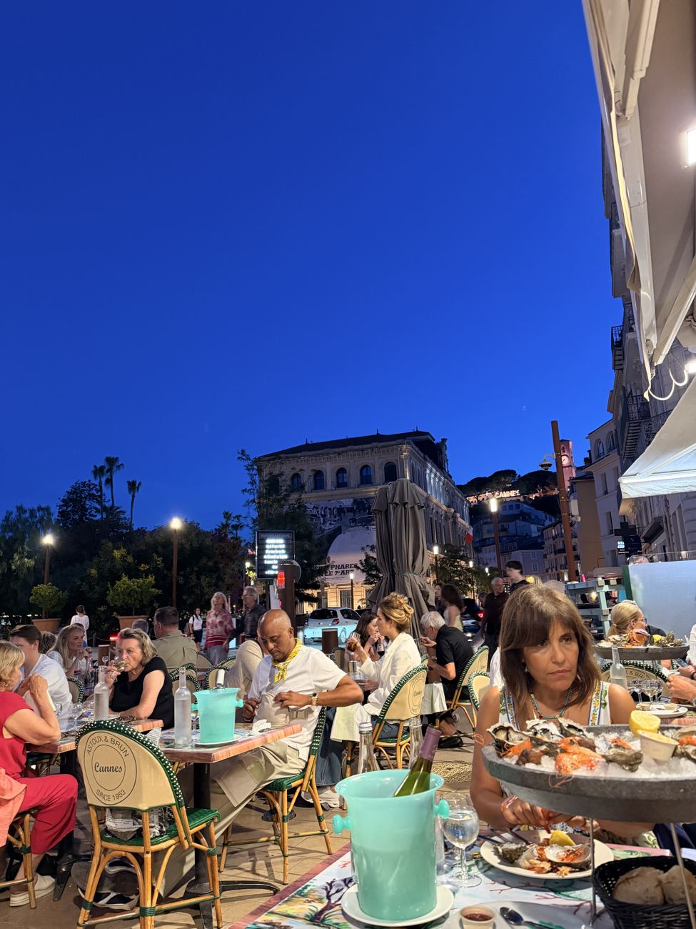 A packed Cannes terrace of cane chairs at blue hour, lamps lit, crowd seated