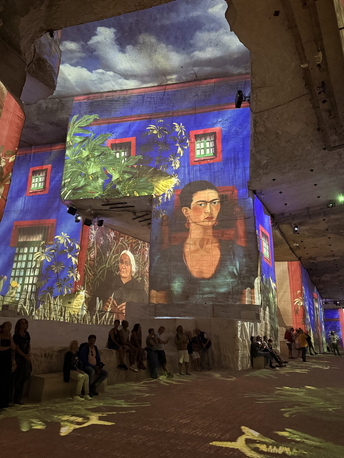 A vast quarry chamber with sky and portraits projected across the walls, visitors small along the floor