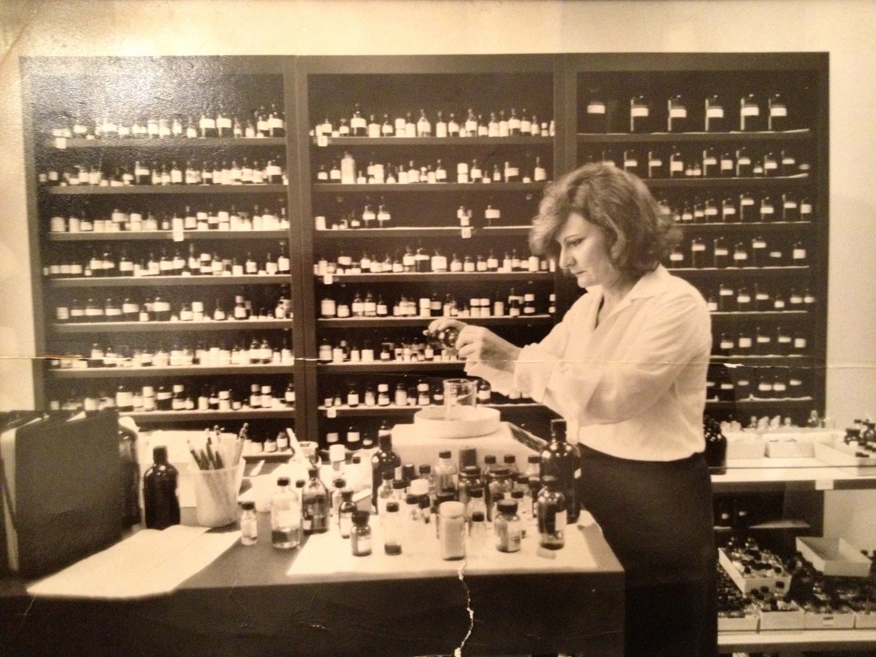 Maman as a young woman at a perfumer's organ, a tiered wall of hundreds of labelled bottles behind her