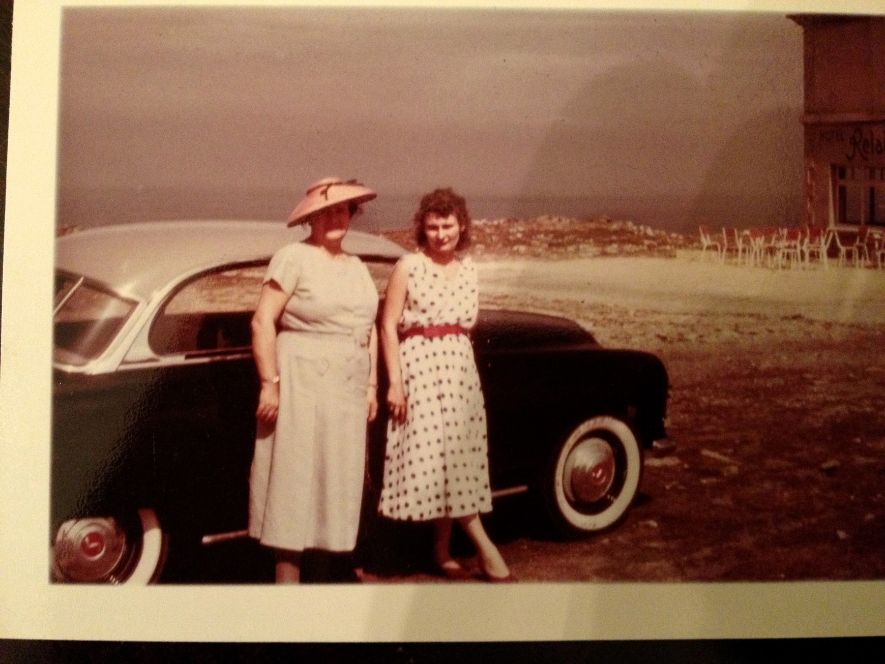 Two women in front of a 1950s car on a stony coastal road