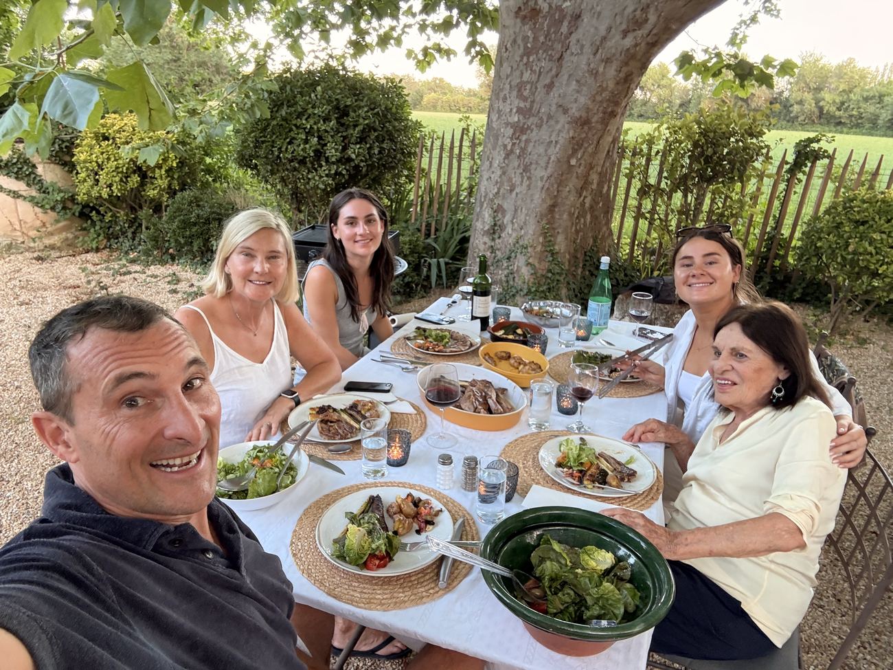 The family around the long table under the trees, plates in front of them