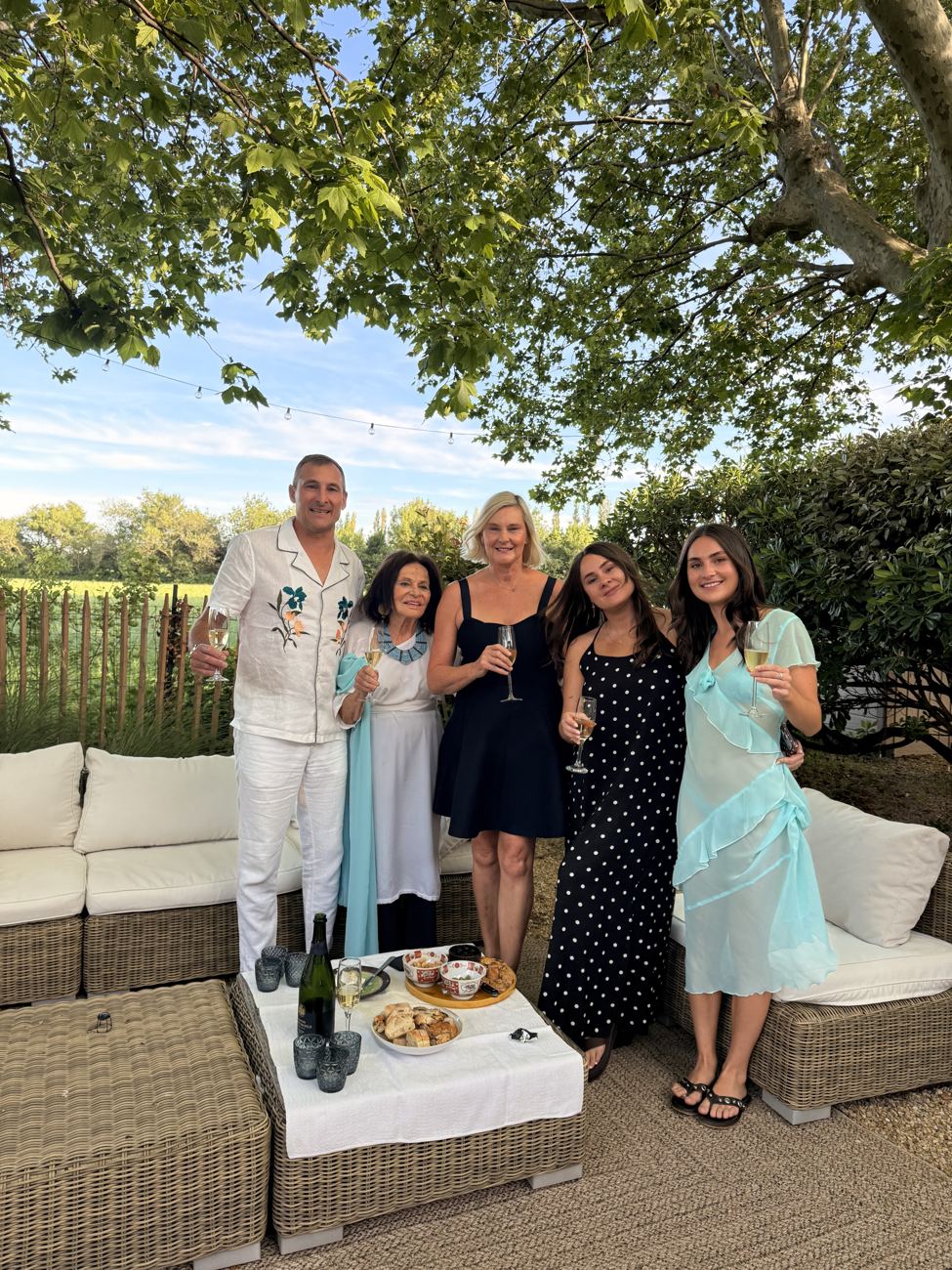 The whole group gathered by the white garden sofas with champagne, golden hour